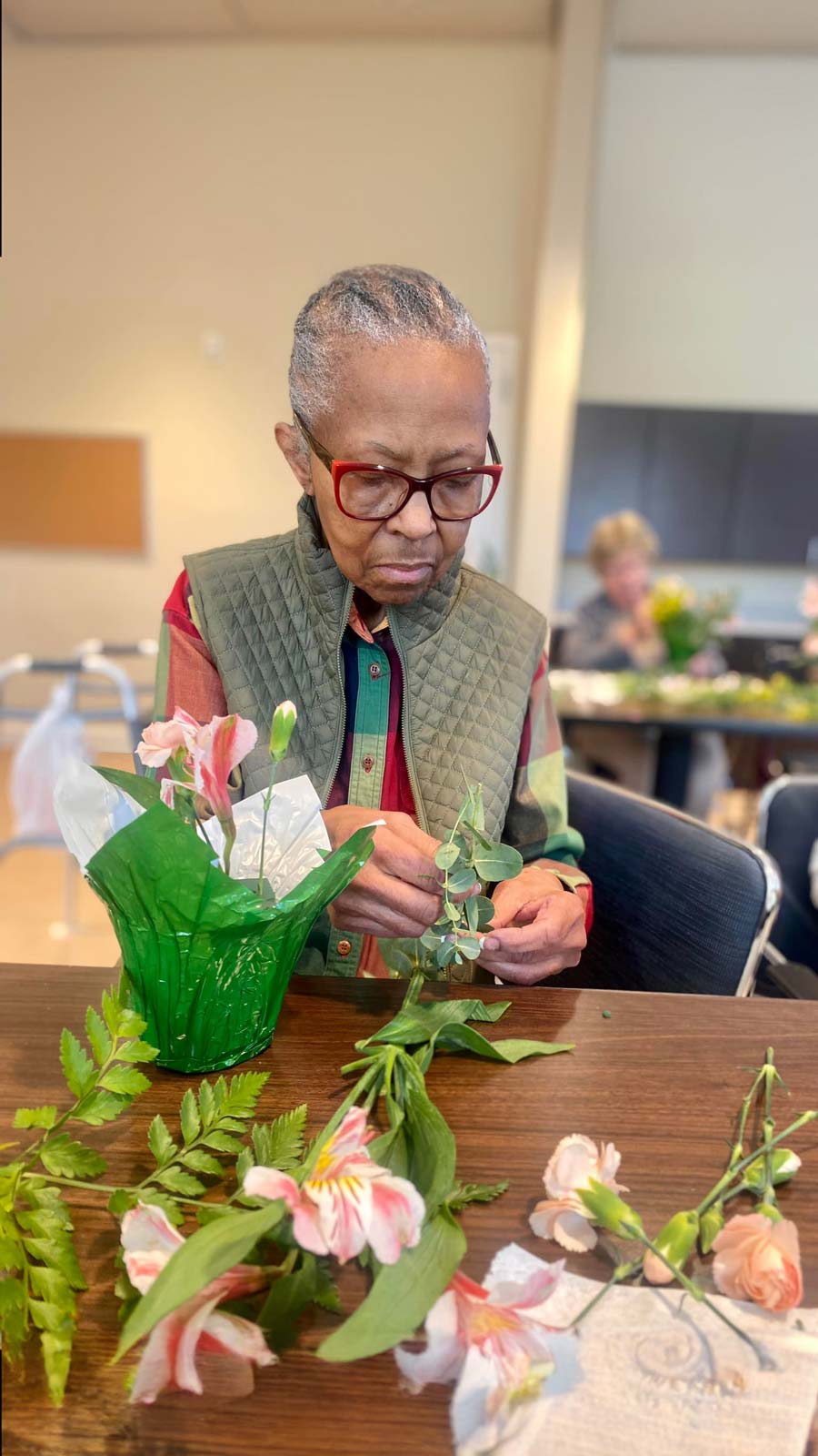 Elderly woman with red glasses concentrating on arranging flowers, enjoying a quiet and creative activity at a table.