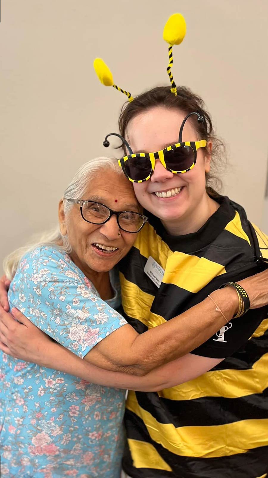 Elderly woman in a blue floral dress joyfully hugging a young woman dressed in a bee costume, both smiling brightly.