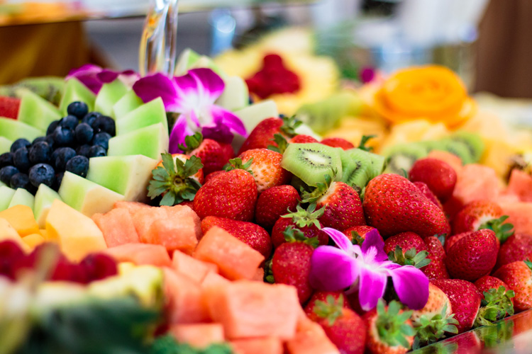 A vibrant and colorful fruit platter featuring an array of fresh strawberries, kiwi slices, watermelon chunks, cantaloupe, honeydew, blueberries, and garnished with decorative flowers, showcasing a delightful and healthy dining option at a senior living community restaurant.