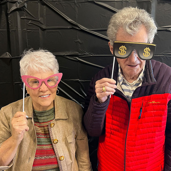 Residents at the senior living community having fun with quirky photo booth props, including oversized dollar sign sunglasses and pink cat-eye glasses, bringing smiles and laughter to their day.