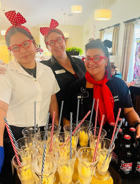 Senior living community staff in retro outfits, ready to serve root beer floats with striped straws, creating a fun nostalgic atmosphere.