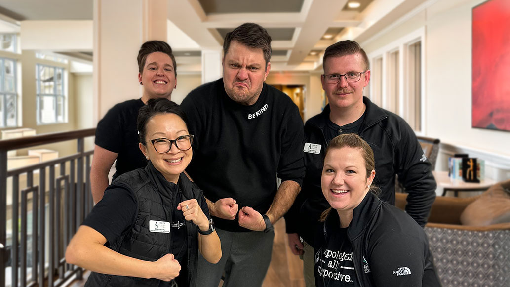 Arrow Senior Living team of five posing playfully in a hallway, showing their muscles and determination during a spring cleaning session.