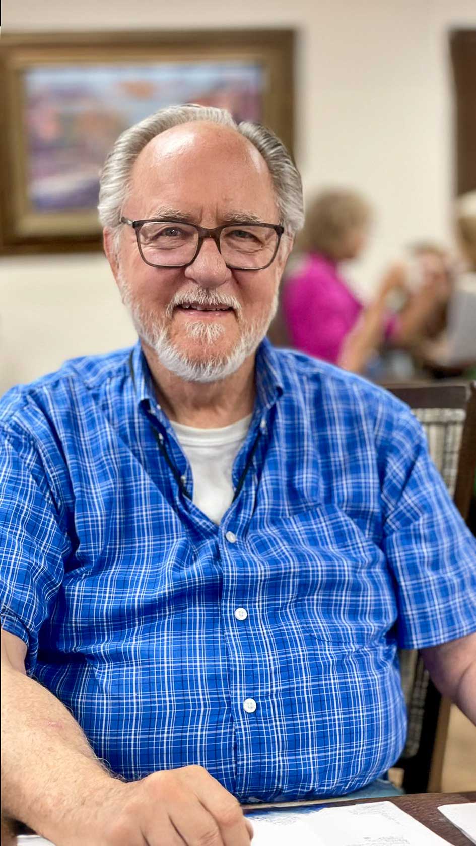 An elderly man in a blue plaid shirt and glasses smiles warmly at the camera, enjoying the vibrant and welcoming atmosphere of the senior living community.