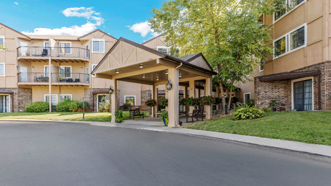 The Cambridge Senior Living facility features a beige exterior with a welcoming covered entrance, surrounded by greenery and benches, providing a warm and inviting atmosphere for residents.