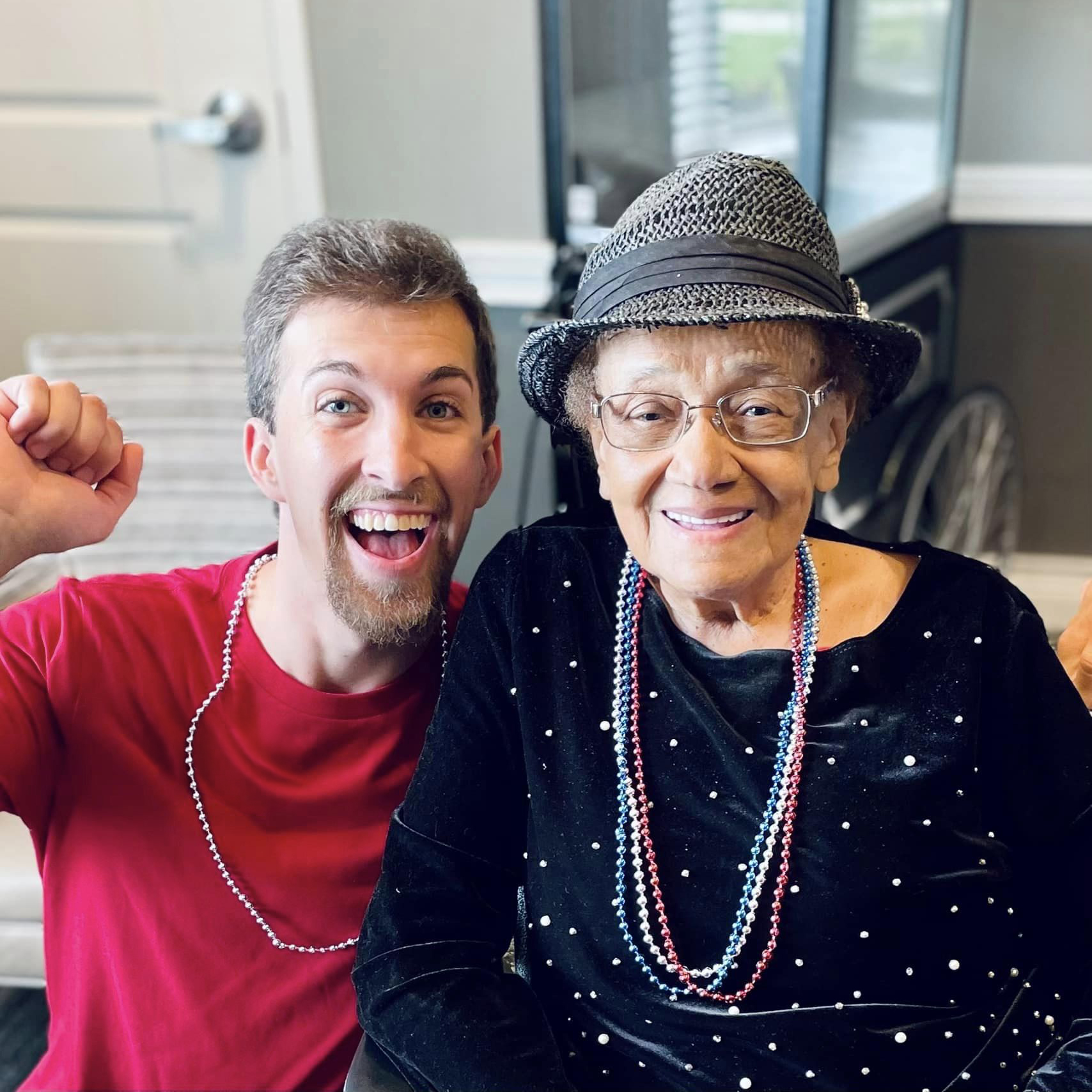 A young man and an elderly woman, both wearing festive beads, smile and celebrate together at a senior living community event.