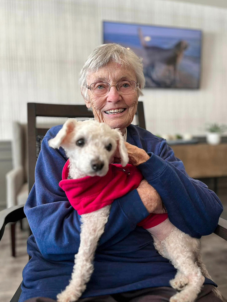 An elderly woman with short gray hair and glasses is smiling while holding a small white dog wearing a red sweater.