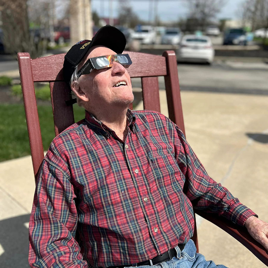 Smiling senior resident wearing a black cap and plaid shirt sits in a rocking chair outdoors, using special eclipse glasses to view a solar eclipse on a sunny day.
