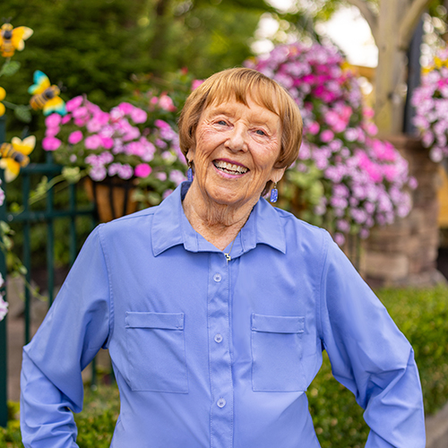 A senior resident wearing a light blue shirt smiles brightly while standing in front of vibrant pink and yellow flowers in a beautifully landscaped outdoor setting.