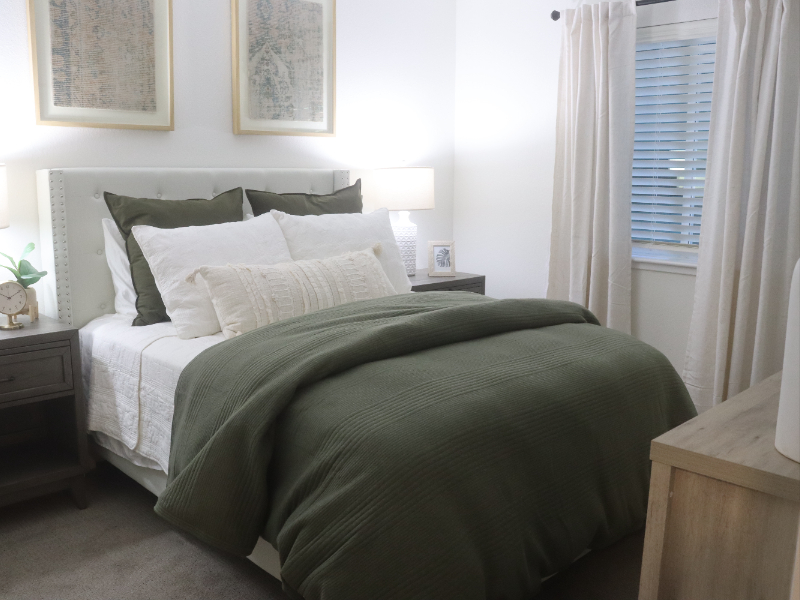 Interior image of a renovated apartment in a senior living community's bedroom area. Bed with white and green bedding, nightstands, white lamps and artwork on the wall.