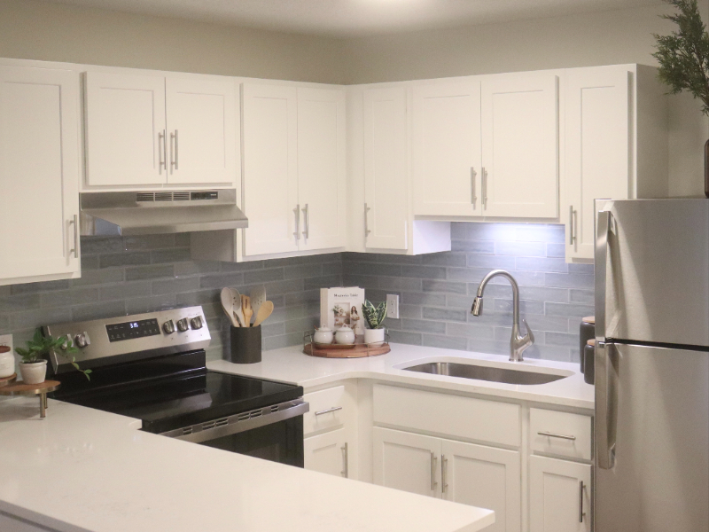 Interior renovated image of the kitchen area of a senior living community apartment. White cabinets and countertops, stainless steel stove, refrigerator, sink and vent hood. Wrap around counter and blue subway backsplash. Decor and plants on countertop.