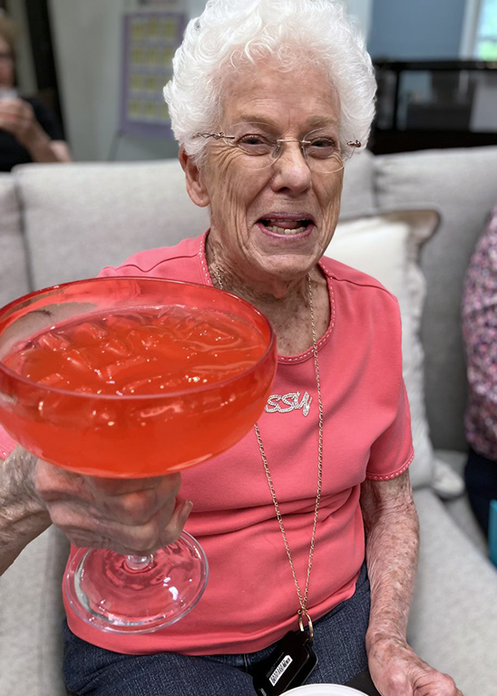 Senior woman in a red shirt excitedly holds up a large icy red margarita glass.
