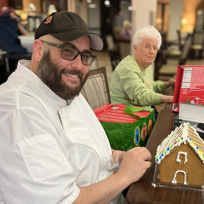 Chef and resident decorating a gingerbread house together at a holiday event, showcasing creativity, teamwork, and festive spirit in a senior living community.