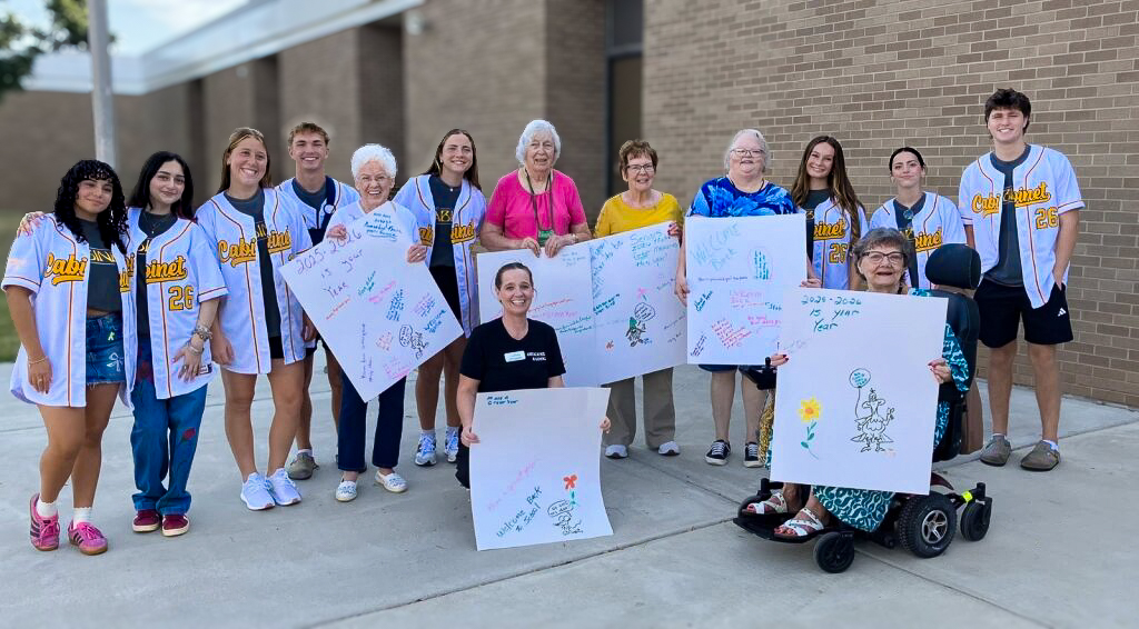 Residents pose with students, holding up handmade signs of encouragement, during an outing to a local first day of school.
