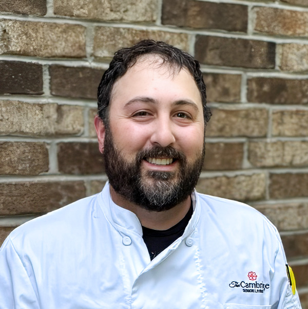 Anthony Smaldino, Culinary Director at Cambridge Senior Living, smiling in a professional headshot with dark curly hair, a full beard, a white chef coat, and a tan and brown brick wall background.