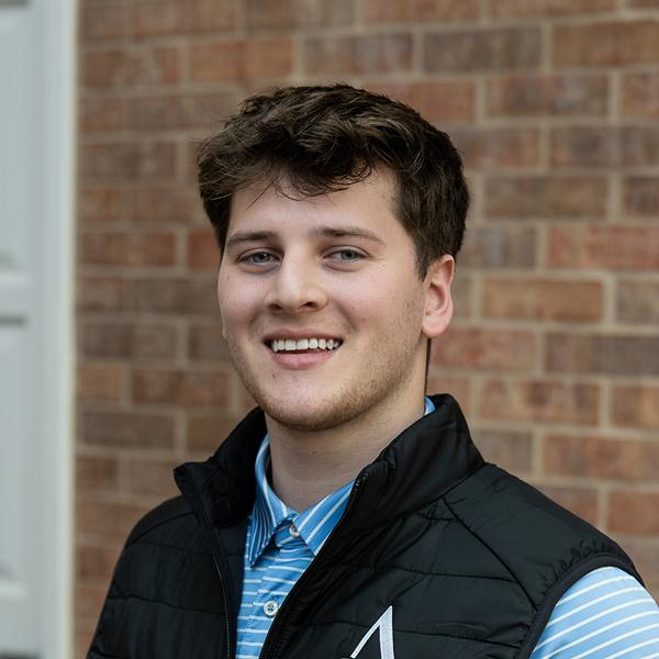Braden McLallen, Plant Operations Director at Cambridge Senior Living, smiling in a professional outdoor headshot with short wavy brown hair, a blue striped collared shirt, a black vest, and a softly blurred brick background.