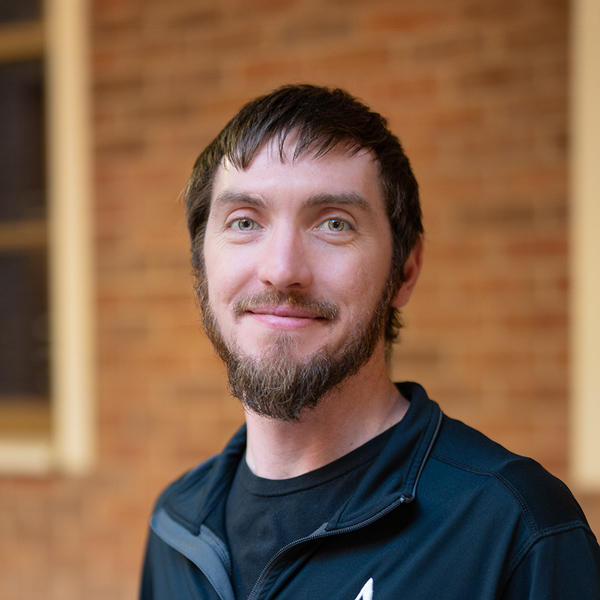 Eric Wells, Business Operations Director at Cambridge Senior Living, smiling in a professional outdoor headshot with short brown hair, a trimmed beard, a black quarter-zip top, and a softly blurred brick background.